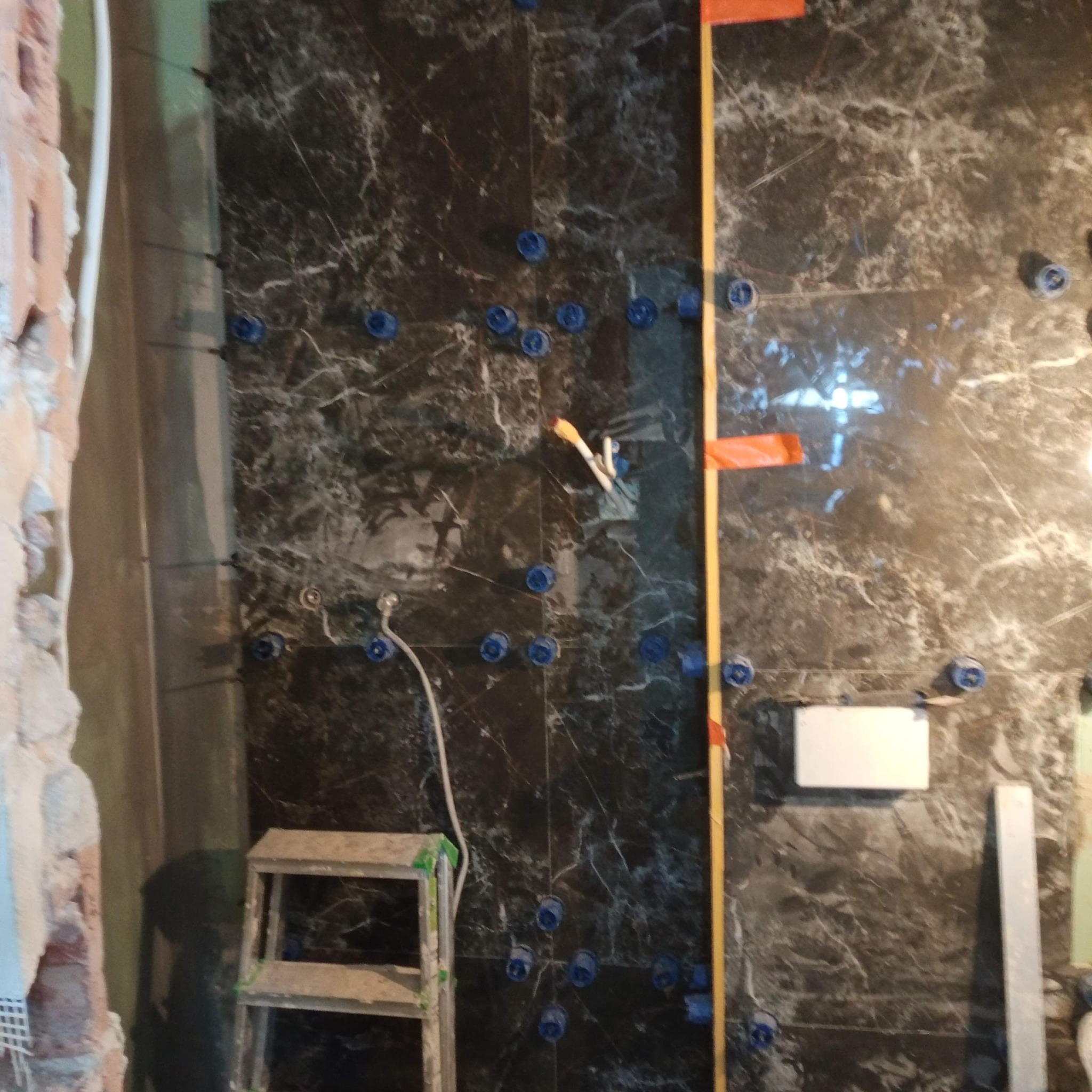 Ciemne płytki imitujące marmur na ścianie w trakcie montażu. Widoczne niebieskie krzyżyki dystansowe, poziomica i fragment instalacji hydraulicznej.