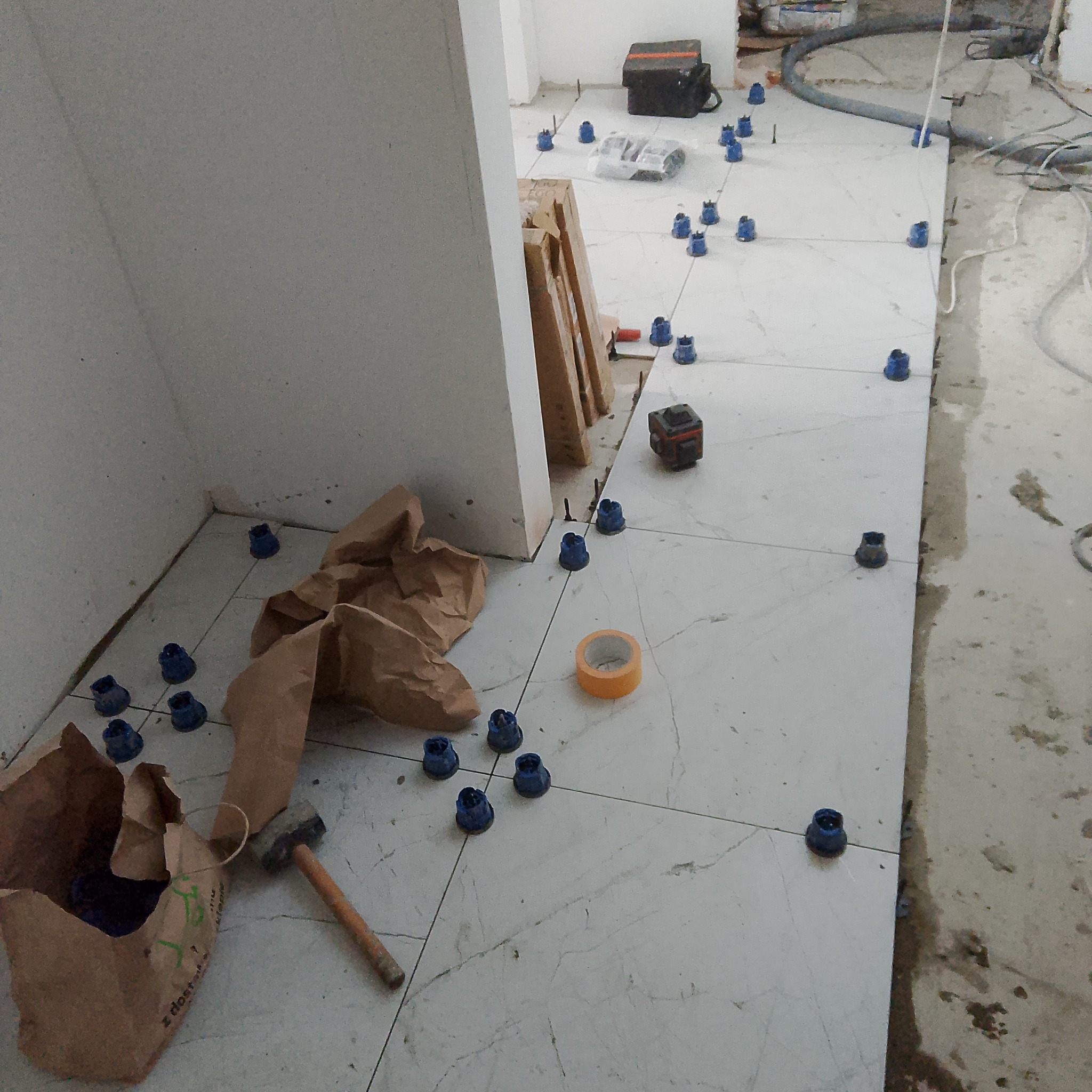 Układanie jasnych płytek podłogowych z użyciem systemu poziomowania, widoczne narzędzia: młotek, poziomica laserowa, taśma. Częściowo wykończone pomieszczenie.