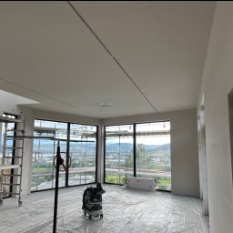 Łukasz Skawski - Wnętrze w stanie deweloperskim z widokiem na miasto, zabezpieczone folią, z widocznym rusztowaniem, szpachlowanymi ścianami i sufitem oraz odkurzaczem przemysłowym.