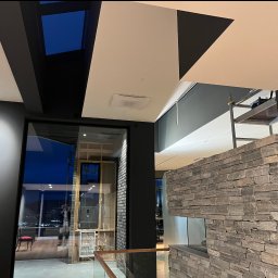 Łukasz Skawski - Nowoczesne wnętrze w trakcie prac wykończeniowych: widoczne fragmenty sufitu z oświetleniem, ściana z kamienia dekoracyjnego, rusztowanie, okno z widokiem na miasto nocą i odblask osoby w oknie.