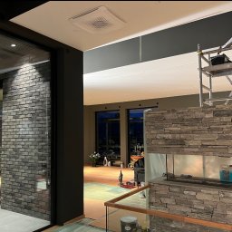 Łukasz Skawski - Mężczyzna na rusztowaniu maluje sufit w nowoczesnym wnętrzu; widoczna ściana z cegły, kamienny kominek i zabezpieczona folią podłoga.