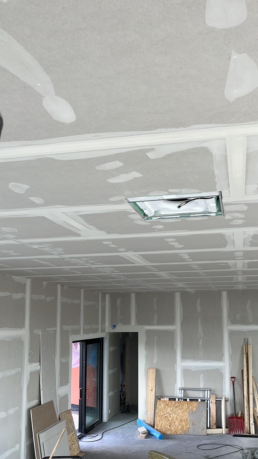 Wnętrze w trakcie prac wykończeniowych, widoczne płyty gipsowo-kartonowe na ścianach i suficie, częściowo zaszpachlowane, z widoczną instalacją oświetleniową w suficie podwieszanym oraz narzędzia...