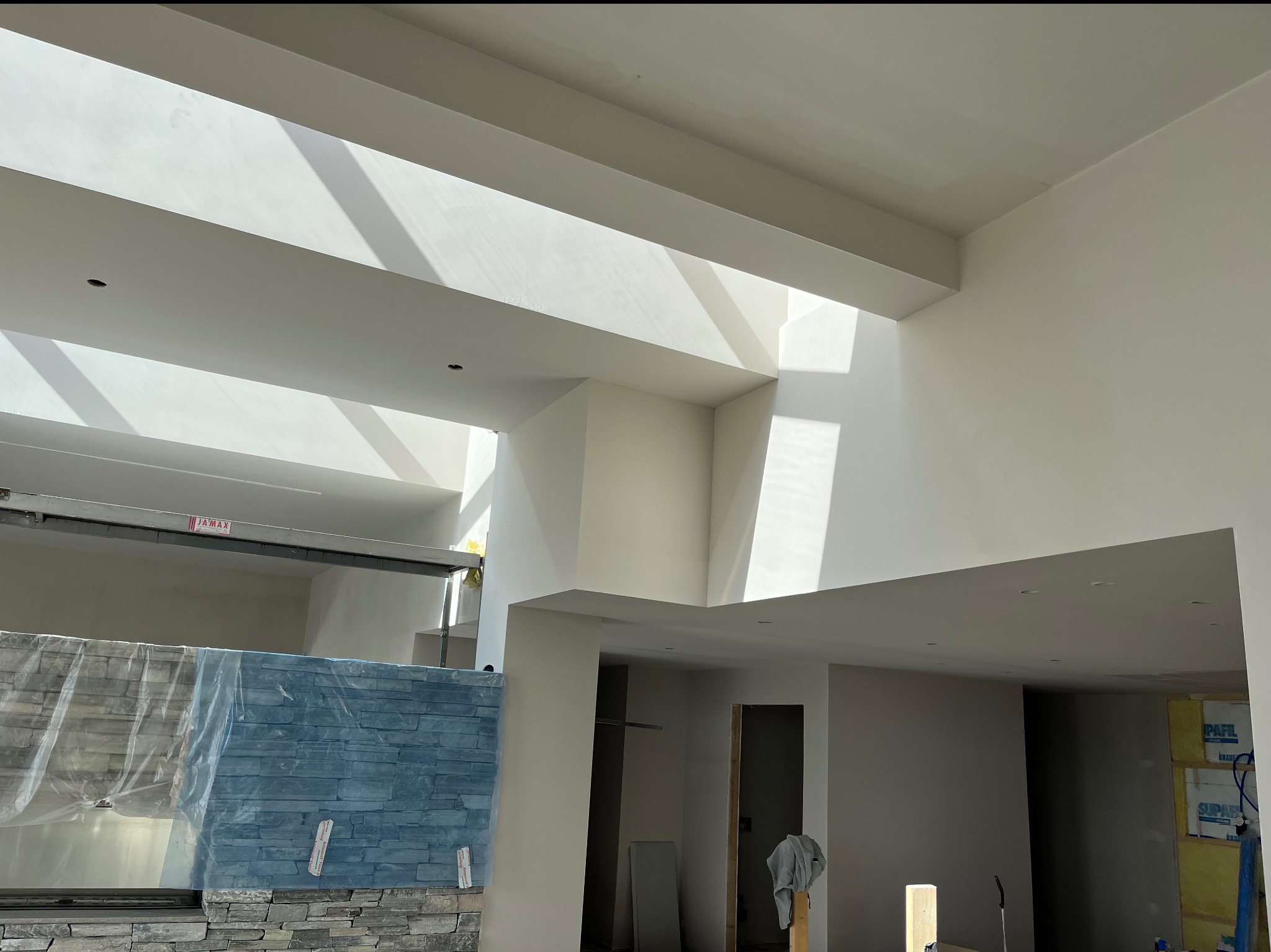 Wnętrze w trakcie wykańczania: pomalowane, jasne ściany i sufit z wbudowanymi punktami świetlnymi, fragment ściany z dekoracyjnym kamieniem częściowo zakryty folią ochronną, widoczne przygotowania...