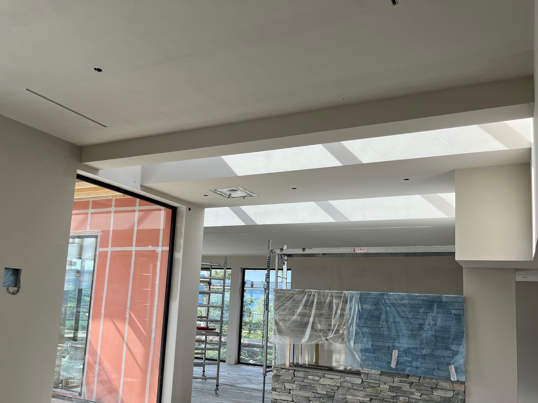 Wnętrze w trakcie prac wykończeniowych: widoczne rusztowanie, zabezpieczona folią ściana z kamienia dekoracyjnego, surowy stan ścian, okno z widokiem na zieleń.