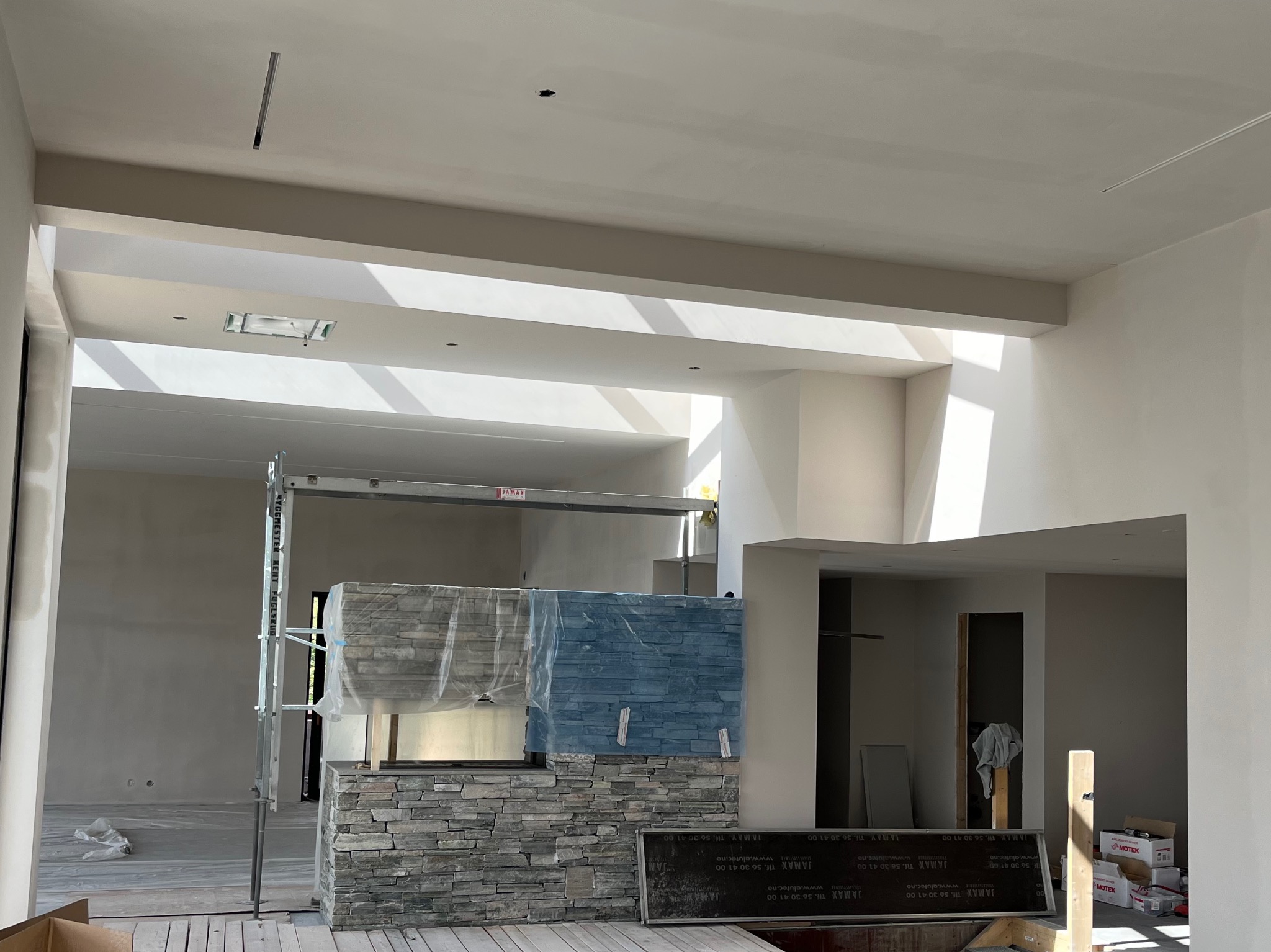 Wnętrze w trakcie prac wykończeniowych z zamontowanym kamieniem dekoracyjnym na ścianie kominkowej, zabezpieczonym folią, widoczne rusztowanie, surowy stan ścian i sufitu przed malowaniem,...