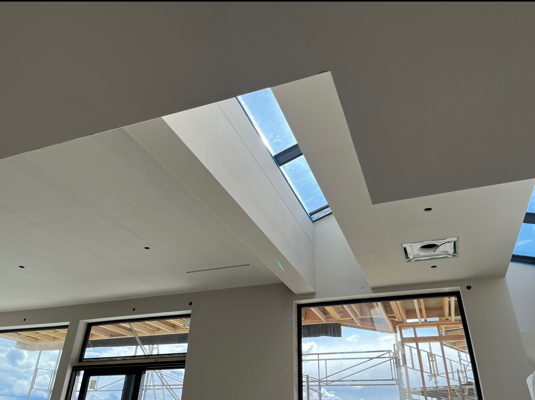 Wnętrze w trakcie prac wykończeniowych: widok na sufit z oknem dachowym i przygotowane otwory pod oświetlenie, ściany w jasnym odcieniu beżu, widoczne rusztowania za oknem.