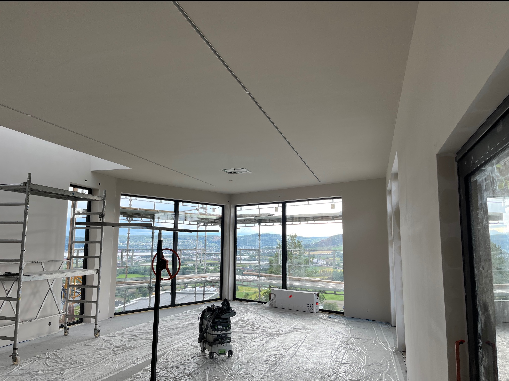 Wnętrze w stanie deweloperskim z widokiem na miasto, zabezpieczone folią, z widocznym rusztowaniem, szpachlowanymi ścianami i sufitem oraz odkurzaczem przemysłowym.