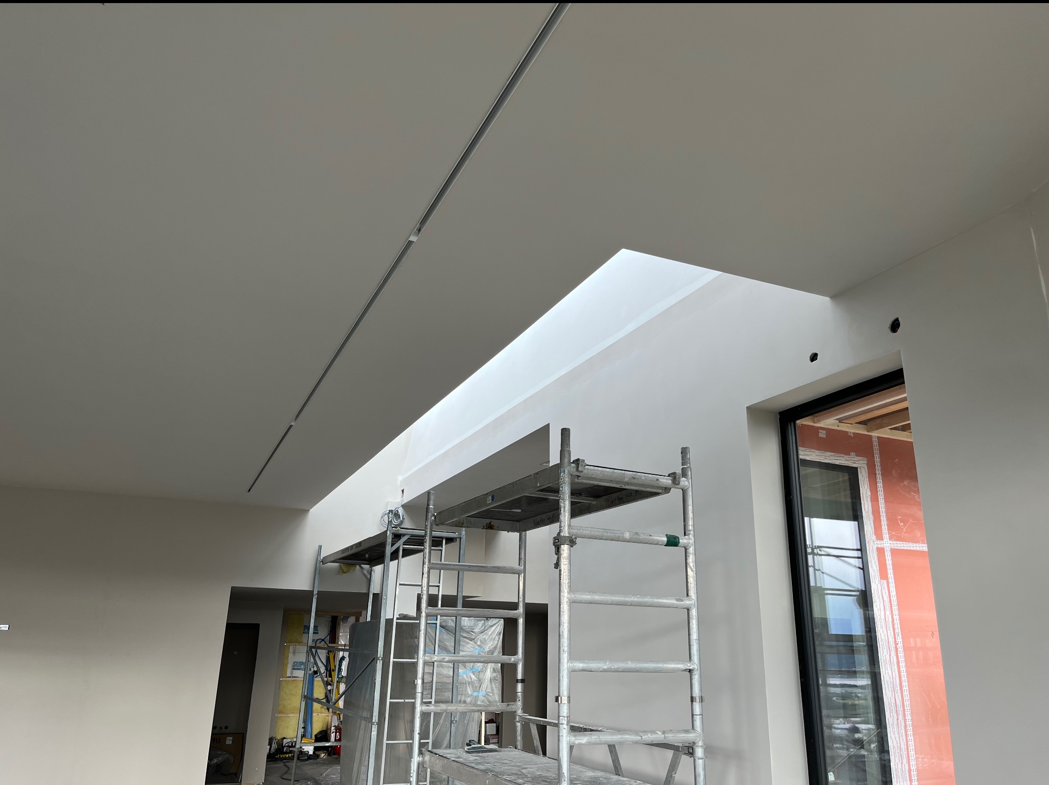 Wnętrze w trakcie prac wykończeniowych: widok na świeżo pomalowany sufit z oknem dachowym, aluminiowe rusztowanie w centrum kadru, ściany przygotowane pod malowanie, widoczne elementy instalacji...