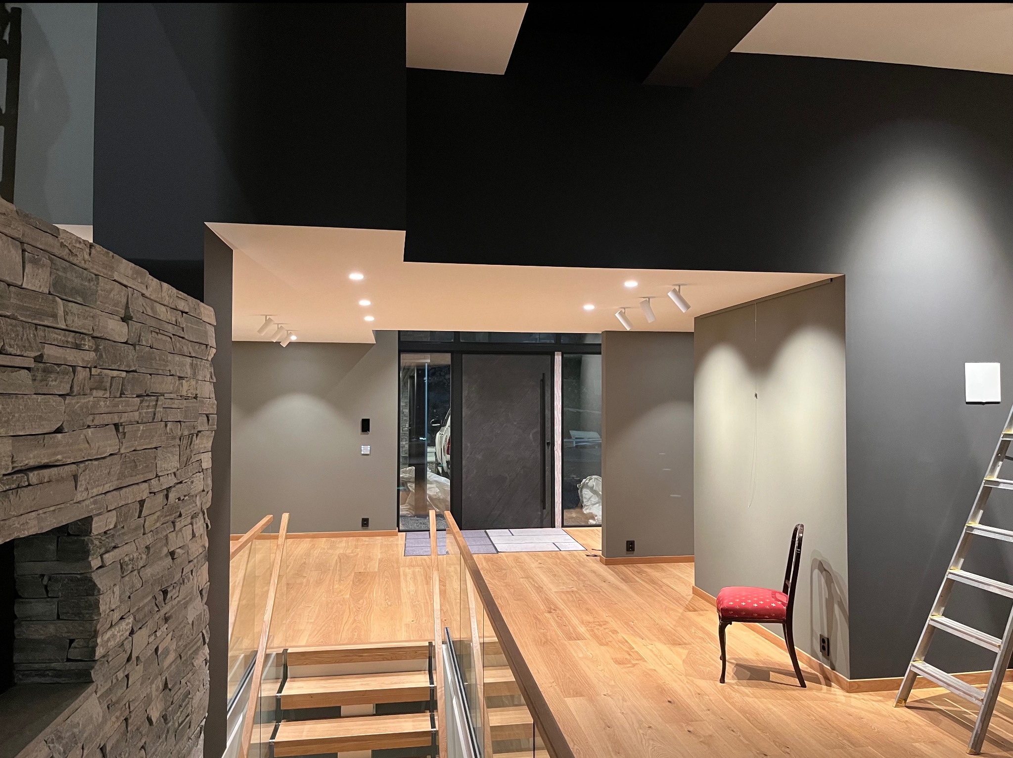 Wnętrze domu w trakcie prac wykończeniowych: pomalowane na szaro ściany, drewniana podłoga, kamienna ściana, schody z drewnianymi stopniami i szklaną balustradą, drabina oparta o ścianę, czerwone...
