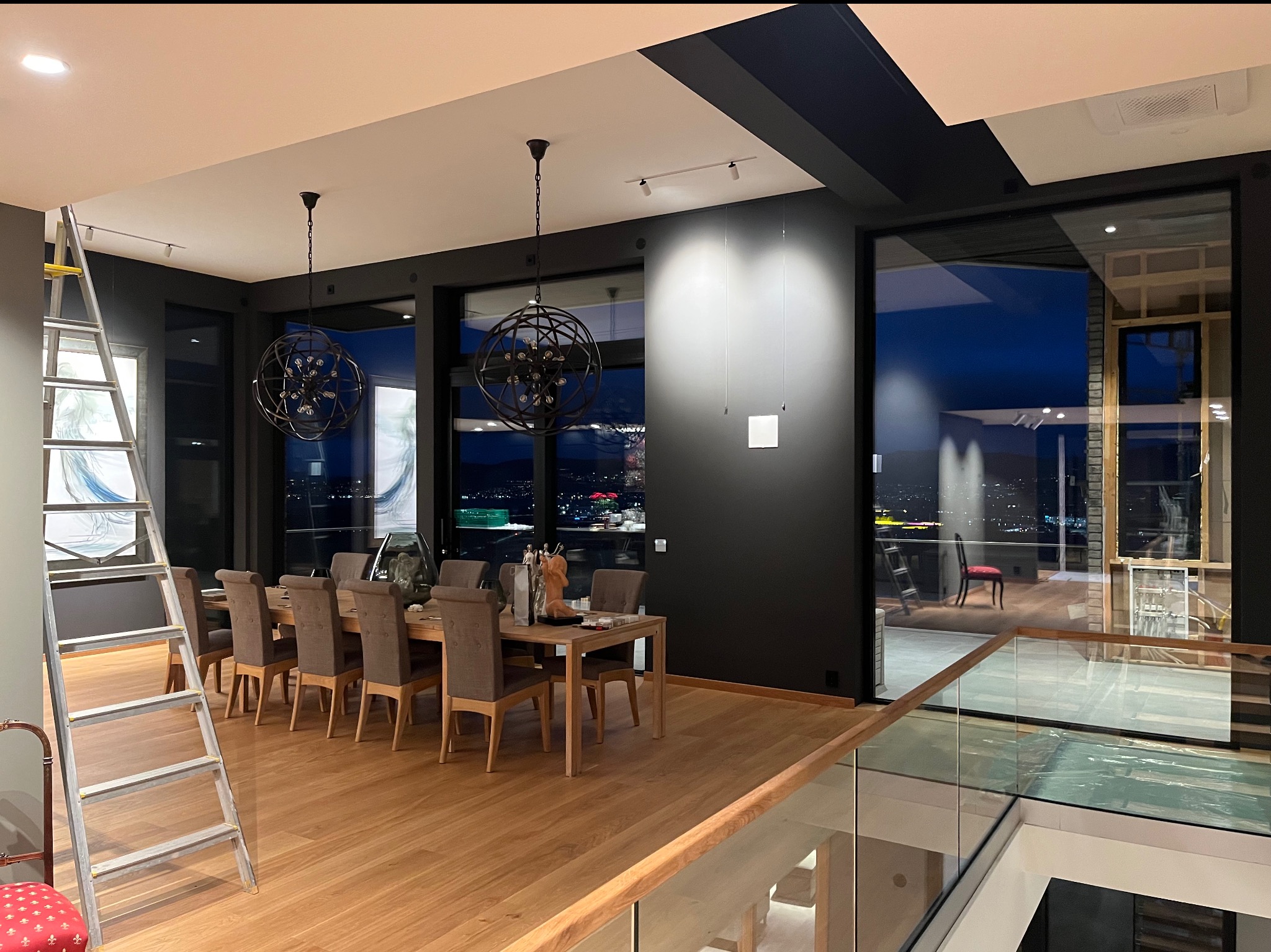 Przestronne wnętrze z drewnianą podłogą i dużym stołem jadalnym, widoczne schody z przezroczystą balustradą, drabina malarska oparta o ścianę, panoramiczne okna z widokiem na miasto w nocy.