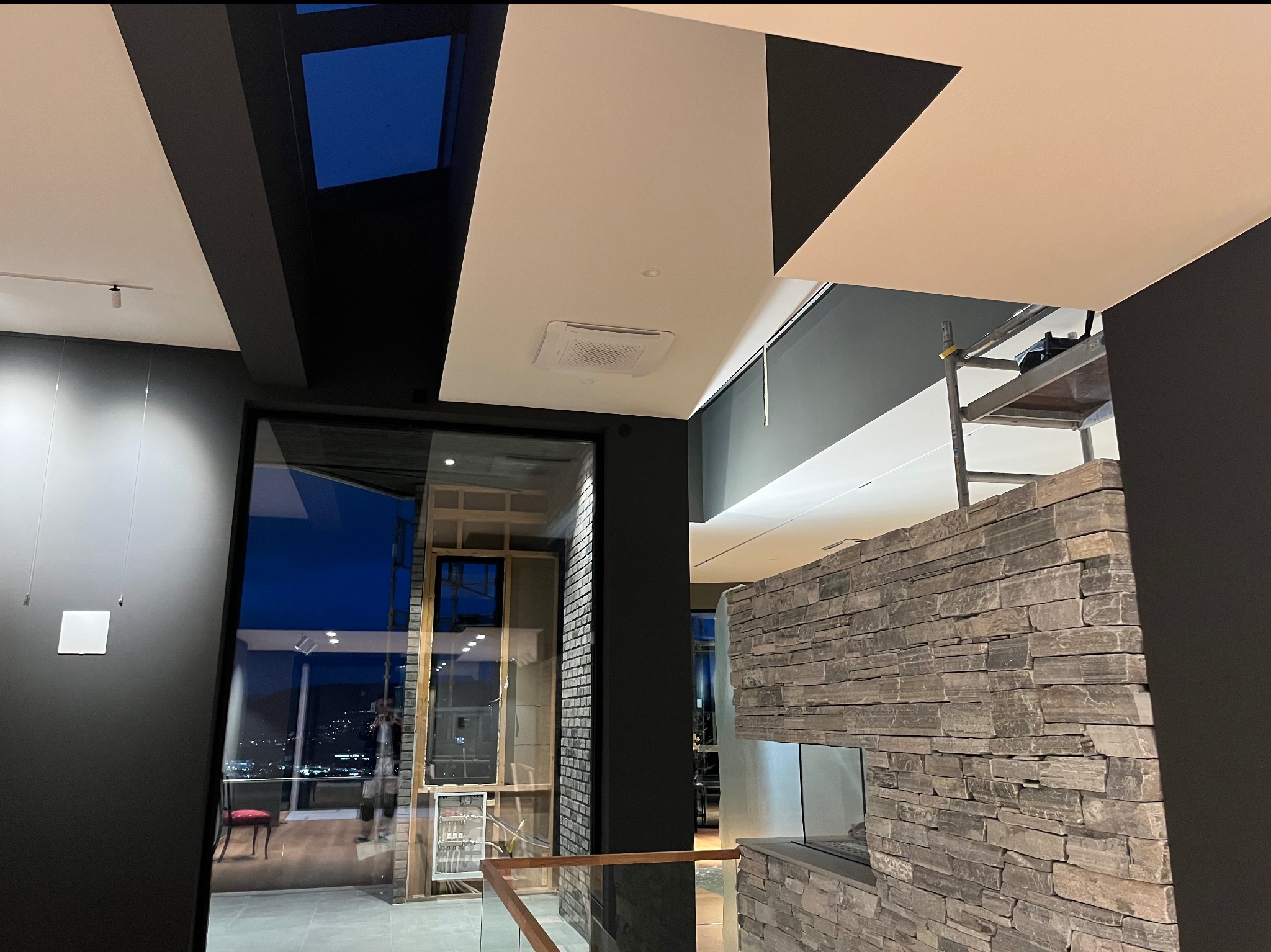 Nowoczesne wnętrze w trakcie prac wykończeniowych: widoczne fragmenty sufitu z oświetleniem, ściana z kamienia dekoracyjnego, rusztowanie, okno z widokiem na miasto nocą i odblask osoby w oknie.