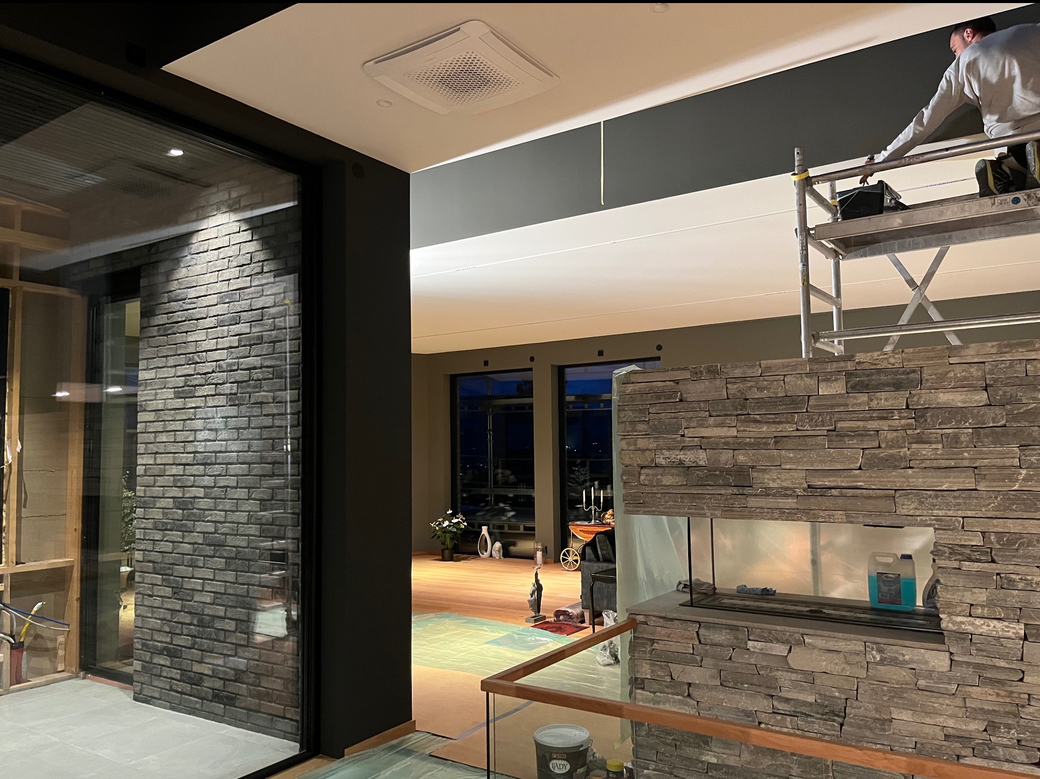 Mężczyzna na rusztowaniu maluje sufit w nowoczesnym wnętrzu; widoczna ściana z cegły, kamienny kominek i zabezpieczona folią podłoga.