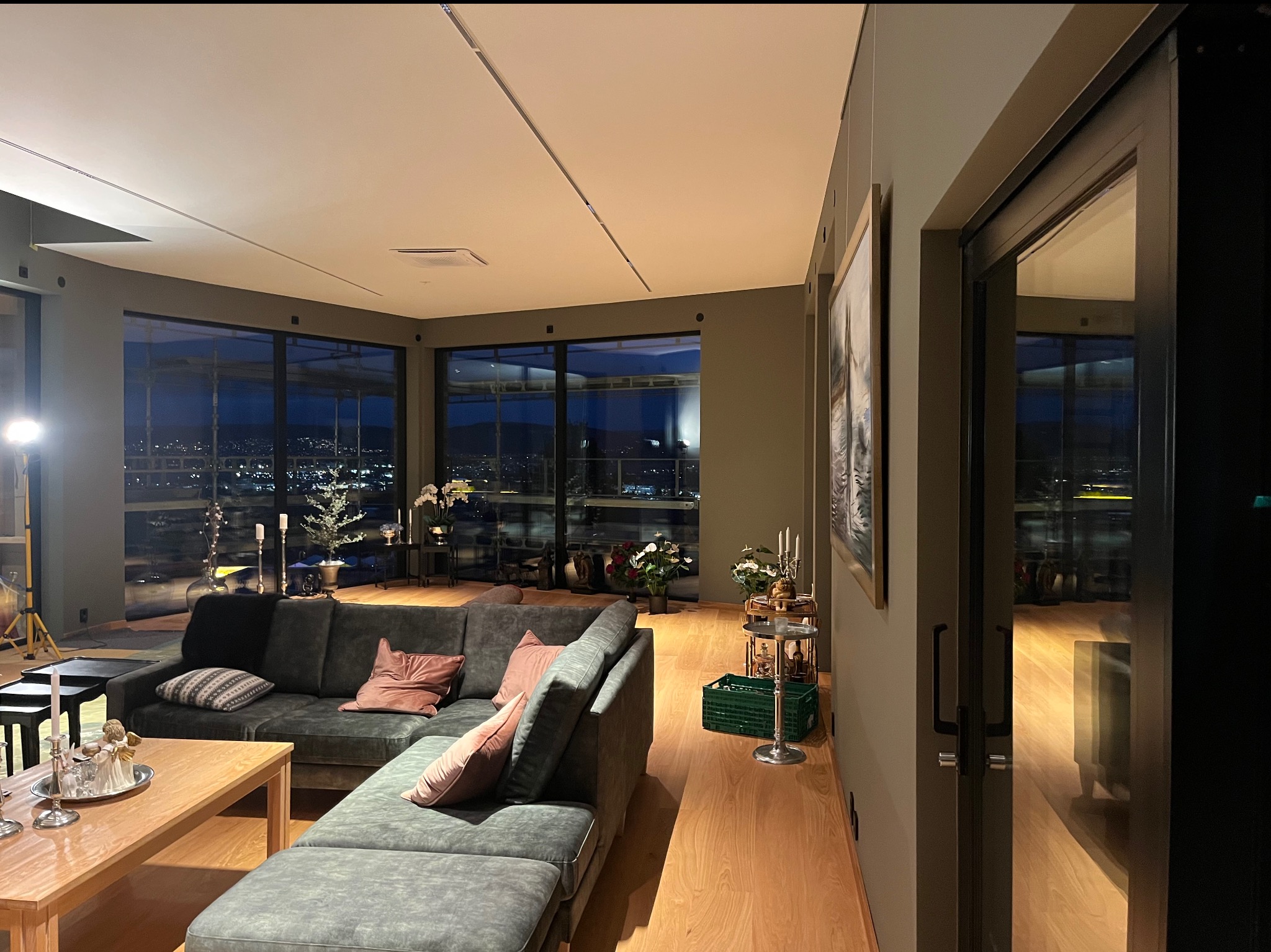 Przestronny salon z dużym narożnikiem w odcieniach szarości, drewnianą podłogą i widokiem na miasto przez panoramiczne okna; minimalistyczne oświetlenie sufitowe i dekoracje.