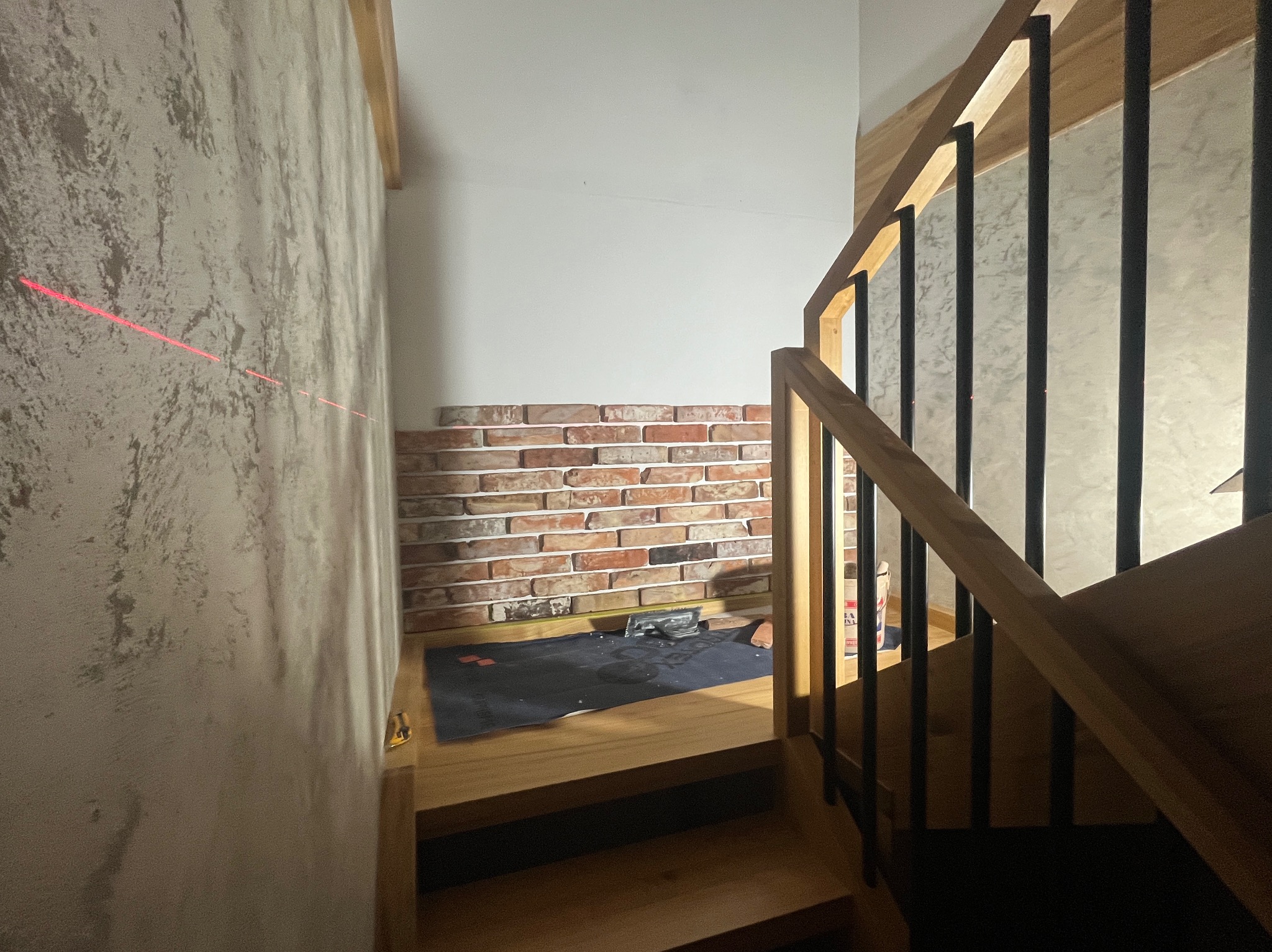 Montaż cegiełek dekoracyjnych na ścianie przy schodach, widoczny laserowy wskaźnik poziomu, narzędzia na podłodze zabezpieczonej folią.