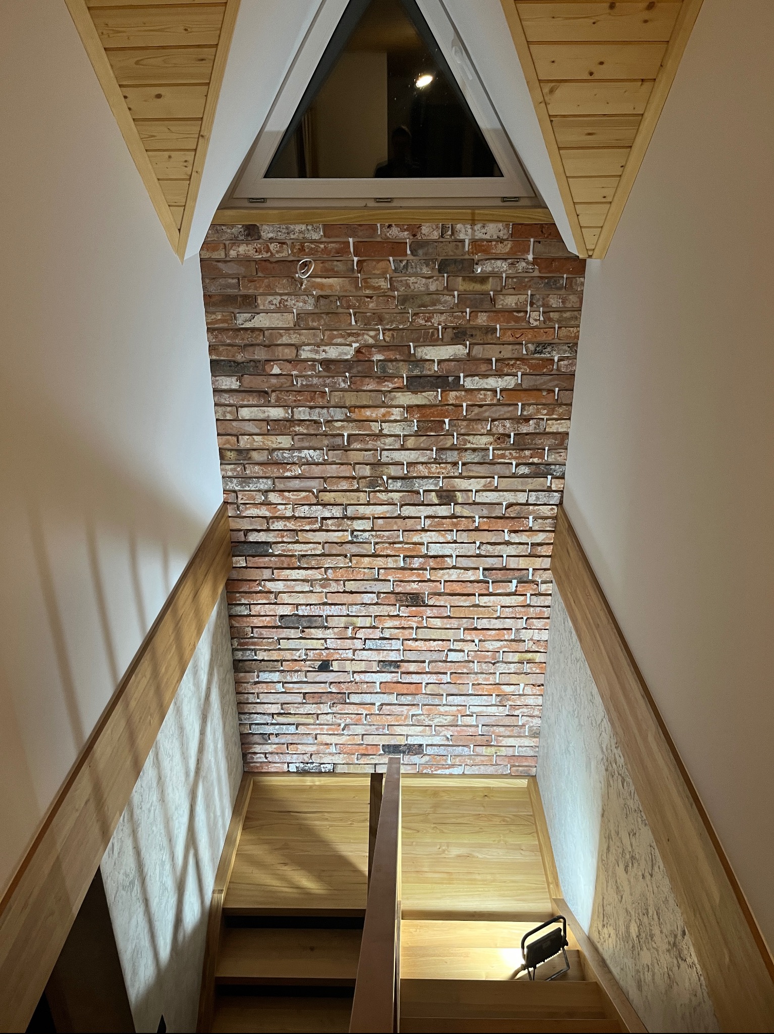 Wnętrze poddasza z drewnianymi schodami, ścianą z cegły klinkierowej pod oknem dachowym i lampą roboczą na schodach.