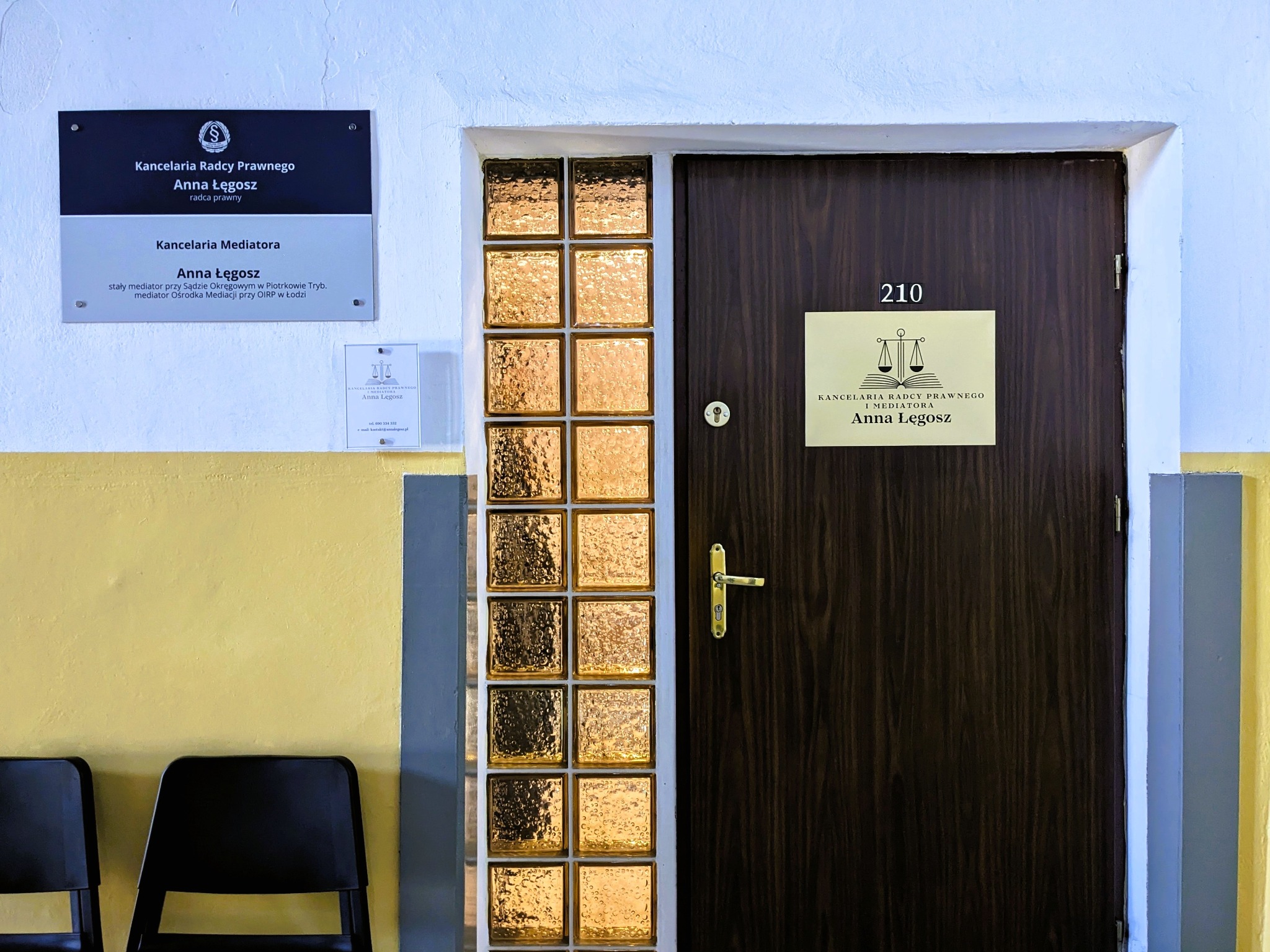 Wejście do kancelarii prawnej: drewniane drzwi z szybami z luksferów, tabliczka z numerem 210 i logo kancelarii Anny Łęgosz, obok tablica informacyjna z tytułem Radca Prawny i Mediator, krzesła...