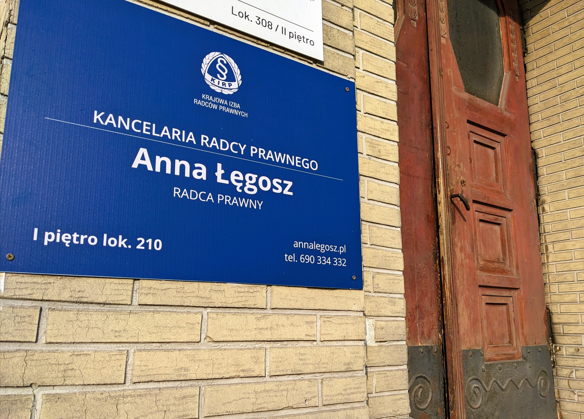 Tablica informacyjna Kancelarii Radcy Prawnego Anny Łęgosz z logo KIRP, umieszczona na ceglanej ścianie obok drewnianych drzwi z dekoracyjnymi elementami.