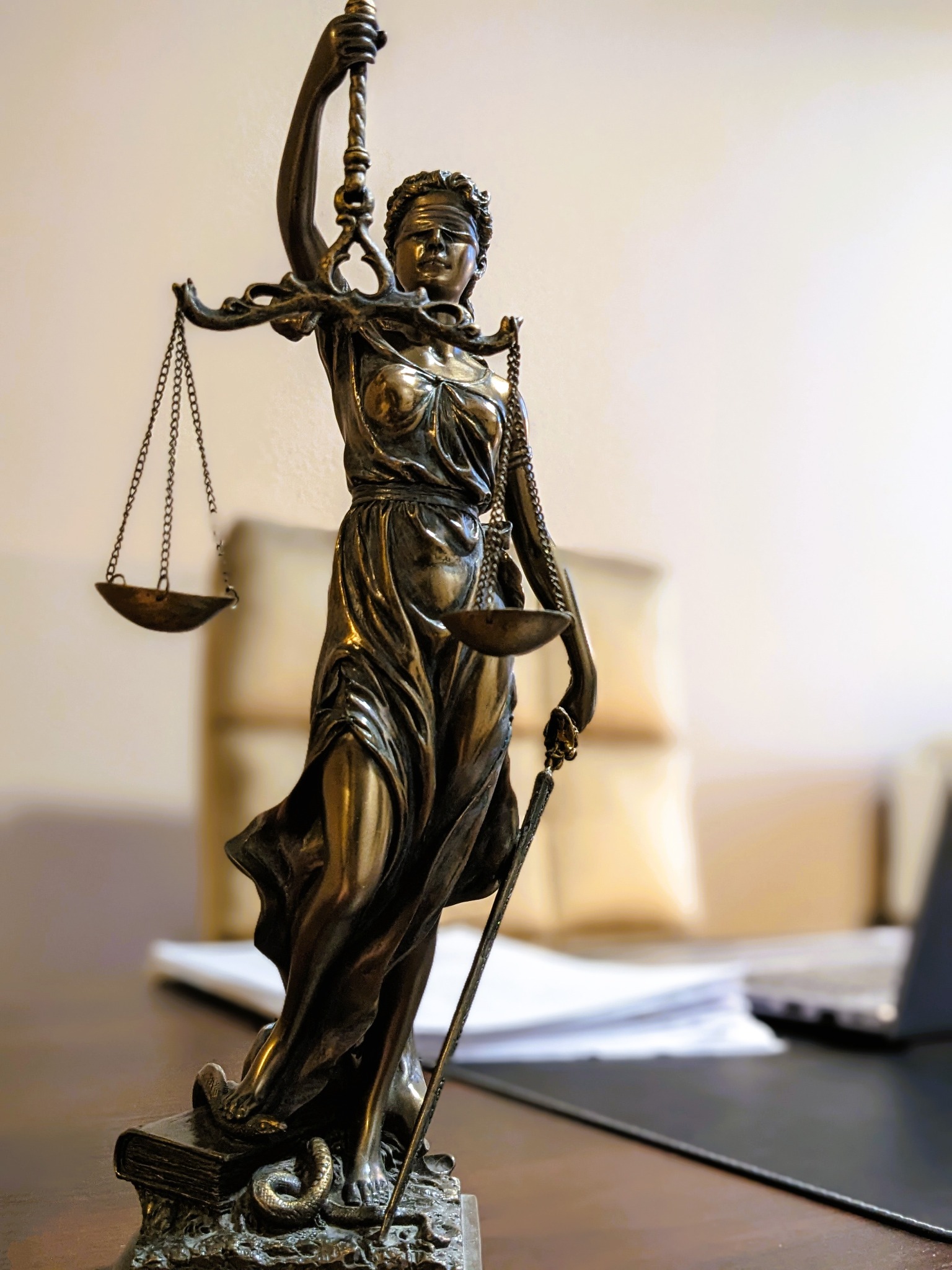 Statuetka Temidy z brązu, bogini sprawiedliwości, z wagą i mieczem, stojąca na książce z wężem, na biurku z dokumentami i laptopem w tle.