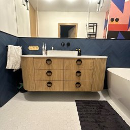 Nookmeble - Nowoczesna łazienka z wiszącą szafką pod umywalkę z drewnianymi frontami i okrągłymi uchwytami, granatowe płytki ułożone w jodełkę na ścianie i biała mozaika na podłodze.