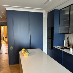 Nookmeble - Nowoczesna kuchnia w odcieniach granatu i bieli, z wyspą o białym blacie i drewnianym wykończeniu, widok na zabudowę kuchenną z piekarnikiem i szafkami, na blacie torba z ziemniakami.