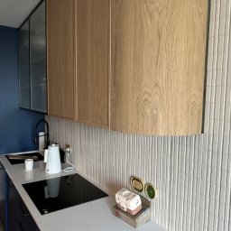 Nookmeble - Nowoczesna kuchnia z górnymi szafkami z jasnego drewna, białym blatem i indukcyjną płytą grzewczą. Na blacie czajnik, kubek i dekoracyjny pojemnik. Tło stanowi ściana z białych, pionowych płytek.