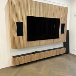 Drewniana zabudowa ściany z telewizorem i głośnikami, z listew w kolorze jasnego drewna, z szafką RTV na dole, parkiet ułożony w jodełkę.