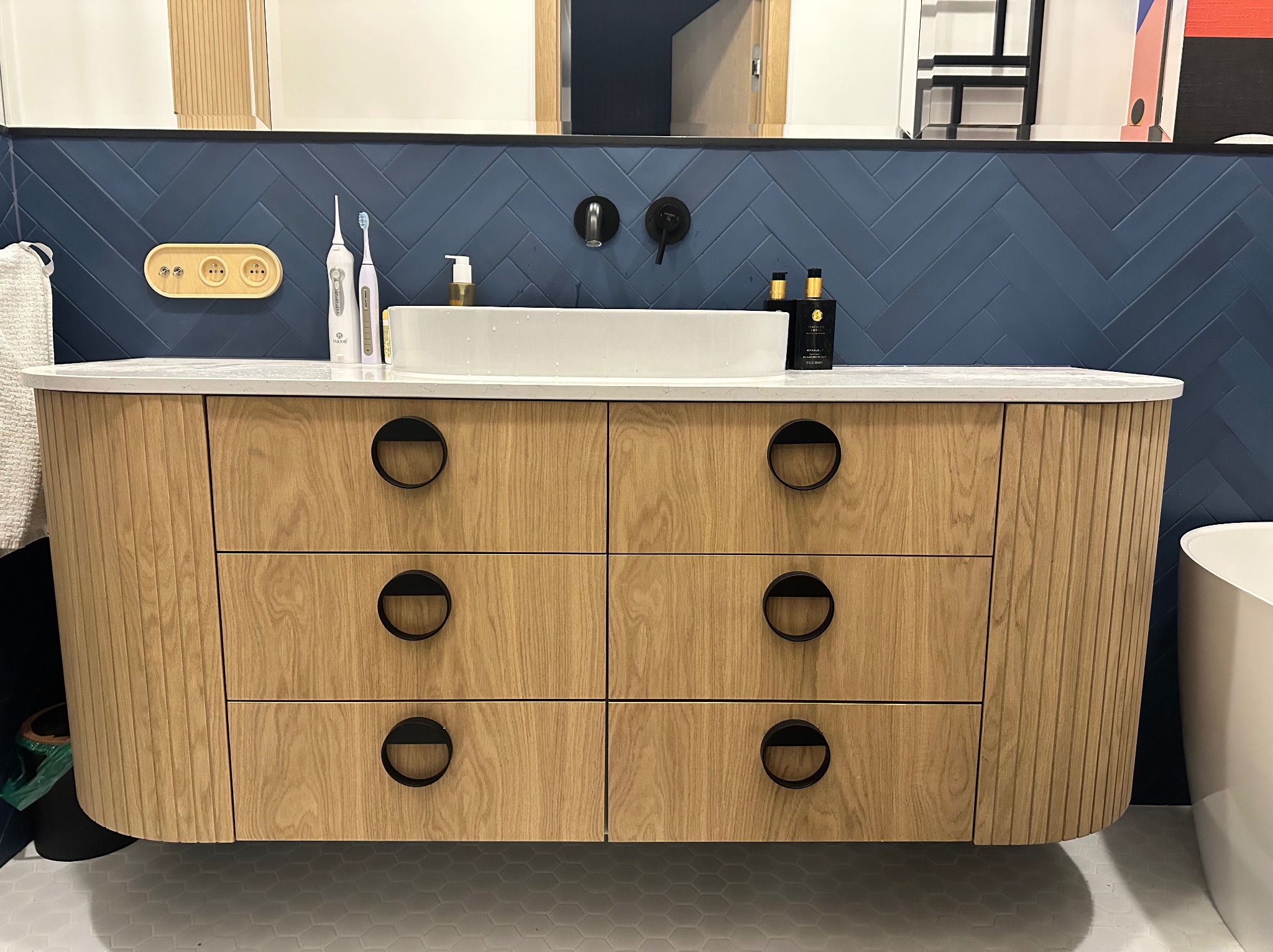 Łazienka z szafką pod umywalkę z drewnianymi frontami szuflad i okrągłymi, czarnymi uchwytami, na tle ściany wyłożonej granatowymi płytkami ułożonymi w jodełkę.