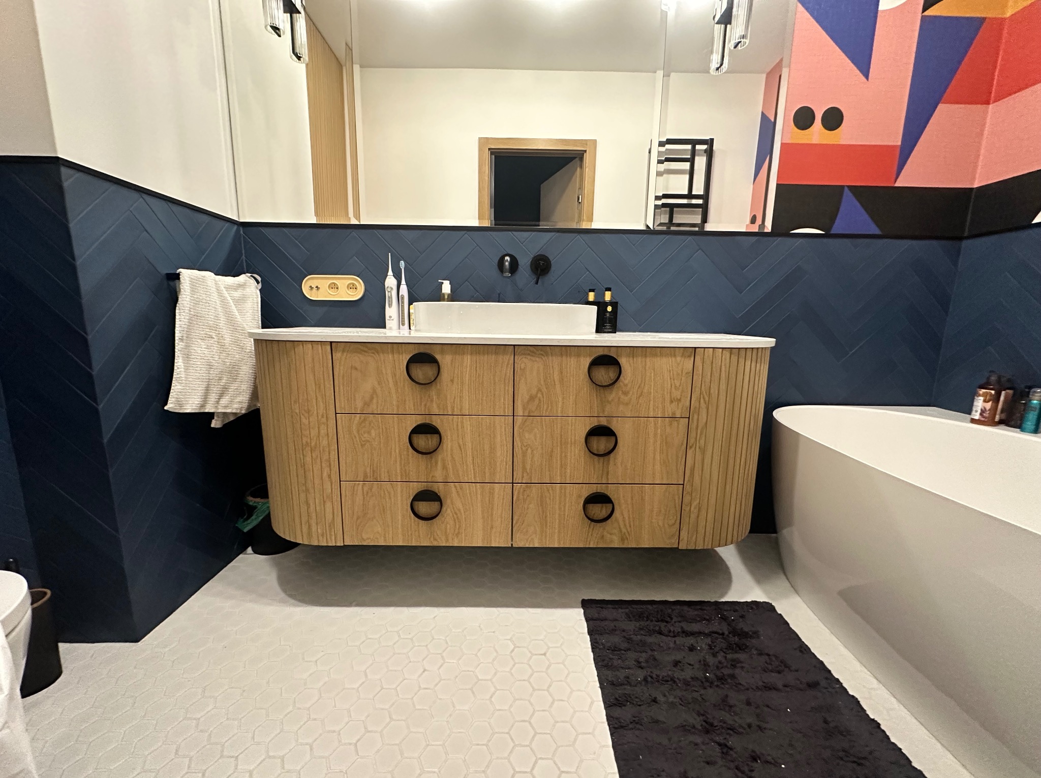 Nowoczesna łazienka z wiszącą szafką pod umywalkę z drewnianymi frontami i okrągłymi uchwytami, granatowe płytki ułożone w jodełkę na ścianie i biała mozaika na podłodze.