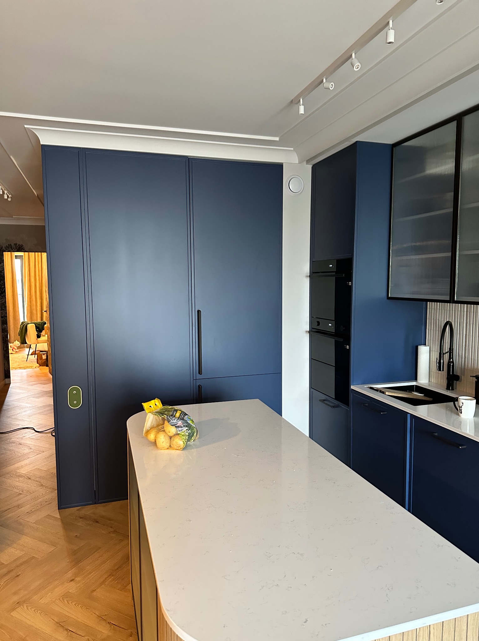 Nowoczesna kuchnia w odcieniach granatu i bieli, z wyspą o białym blacie i drewnianym wykończeniu, widok na zabudowę kuchenną z piekarnikiem i szafkami, na blacie torba z ziemniakami.