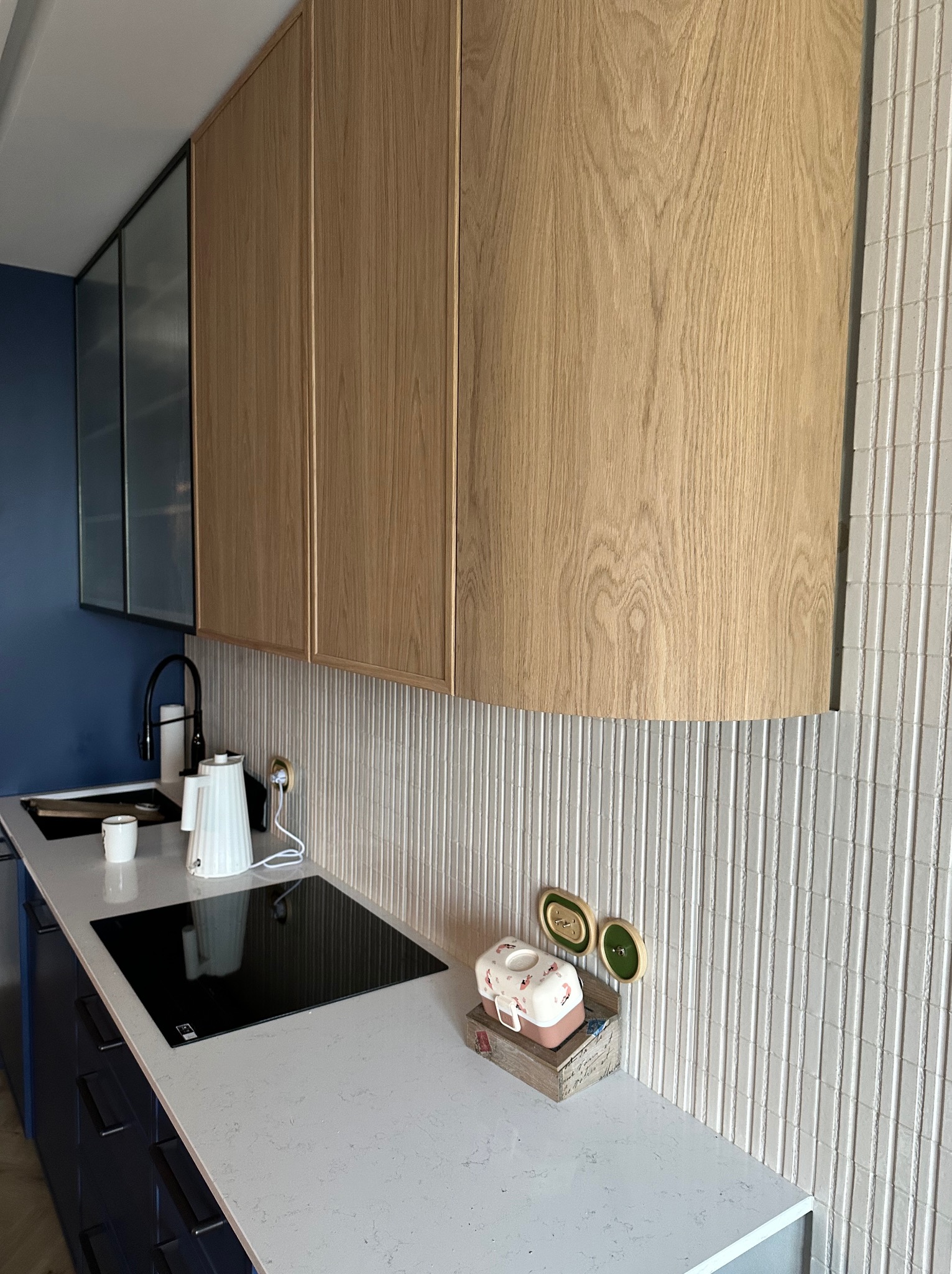 Nowoczesna kuchnia z górnymi szafkami z jasnego drewna, białym blatem i indukcyjną płytą grzewczą. Na blacie czajnik, kubek i dekoracyjny pojemnik. Tło stanowi ściana z białych, pionowych płytek.