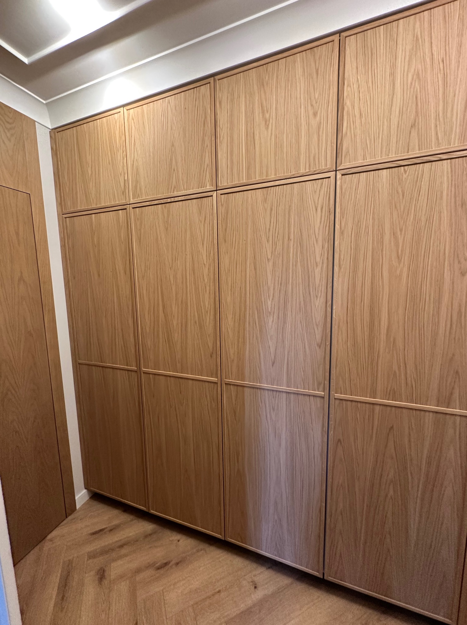 Duża, zabudowana szafa z jasnego drewna z subtelnym wzorem słojów, podzielona na segmenty z delikatnymi, wpuszczanymi ramkami, dopasowana do podłogi w jodełkę.