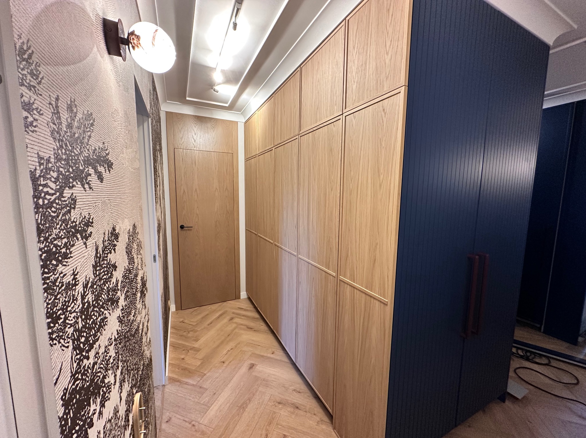 Długa, zabudowana szafa z jasnego drewna z ciemnoniebieskim segmentem, dopasowana do drewnianej podłogi ułożonej w jodełkę i wzorzystej tapety, oświetlona lampą ścienną.