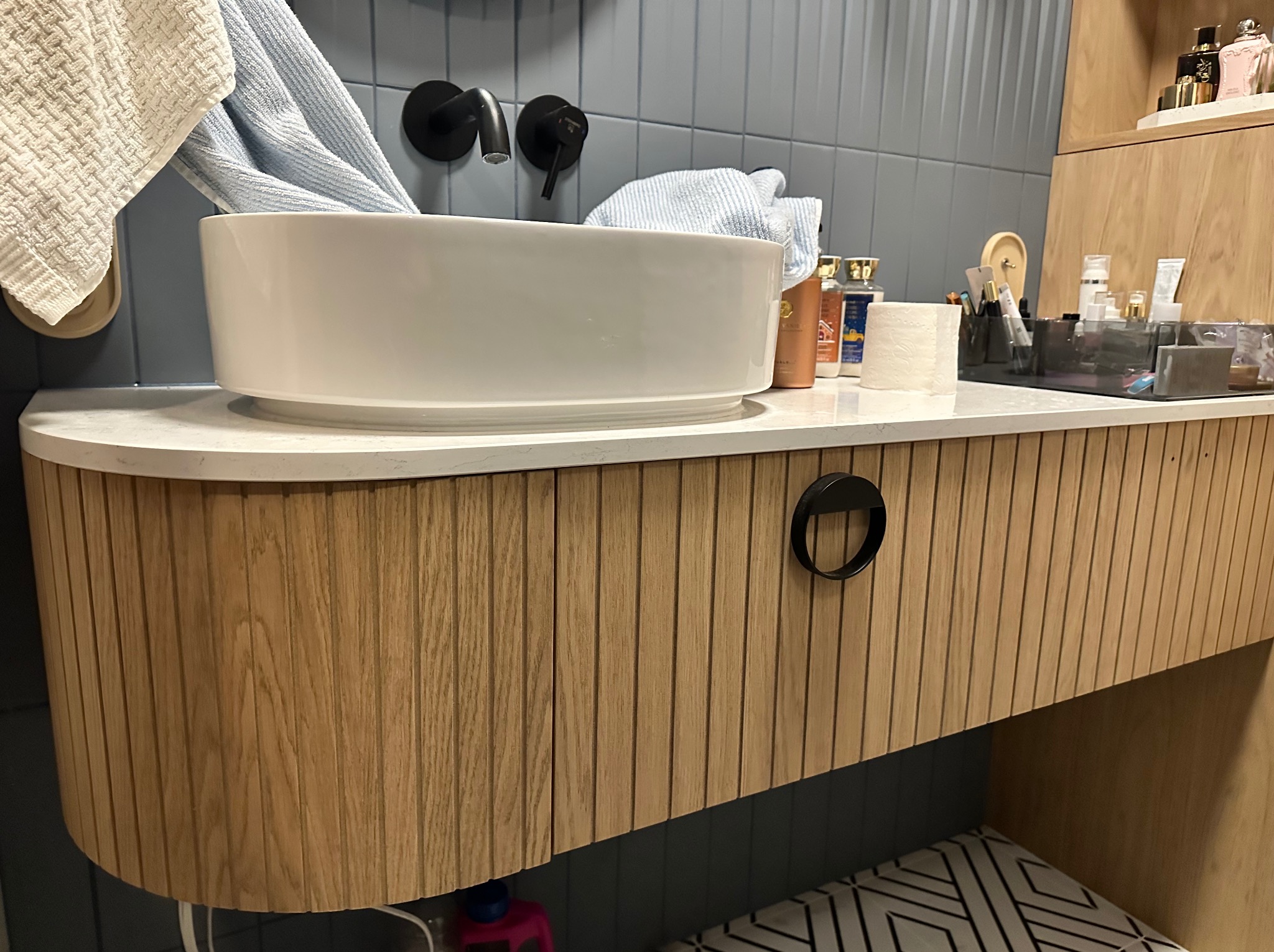 Nowoczesna łazienka z białą umywalką nablatową, czarną armaturą i szafką z drewnianymi lamelami. Na blacie kosmetyki i ręczniki.