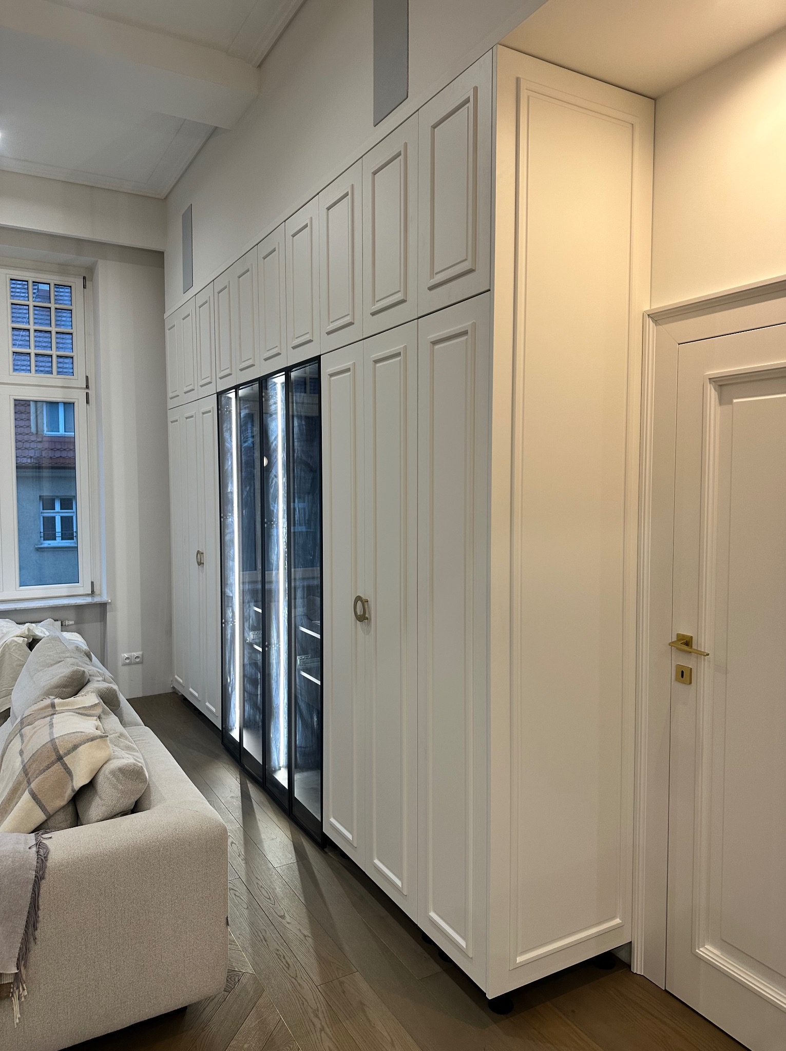 Długa, biała zabudowa meblowa z frezowanymi frontami, w tym przeszklona witryna z oświetleniem wewnętrznym, na tle drewnianej podłogi i fragmentu sofy w jasnym salonie.