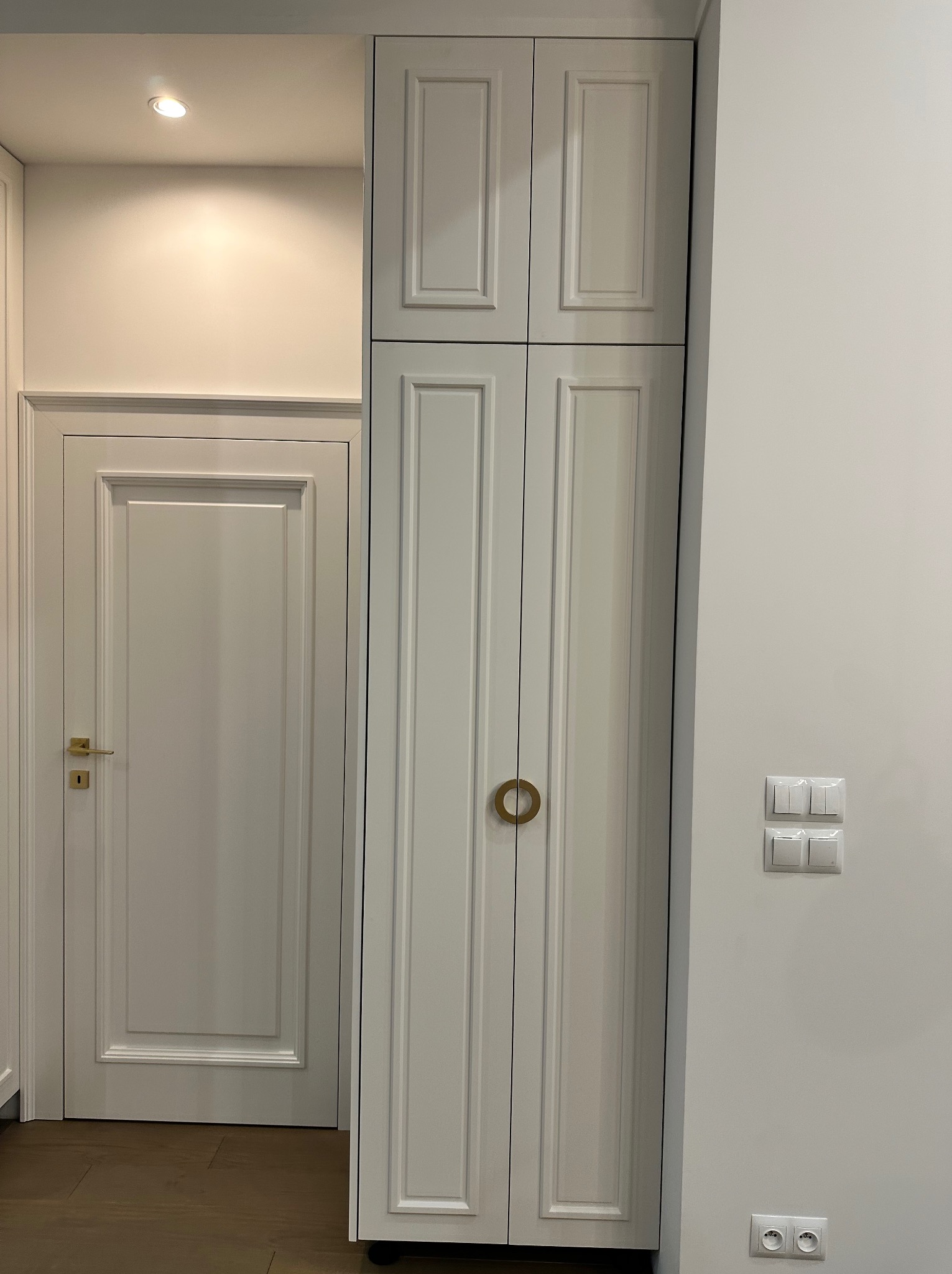 Biała, wysoka szafa z frezowanymi drzwiami i okrągłymi uchwytami, stojąca obok białych drzwi z mosiężną klamką, na tle białej ściany z włącznikami i gniazdkami.