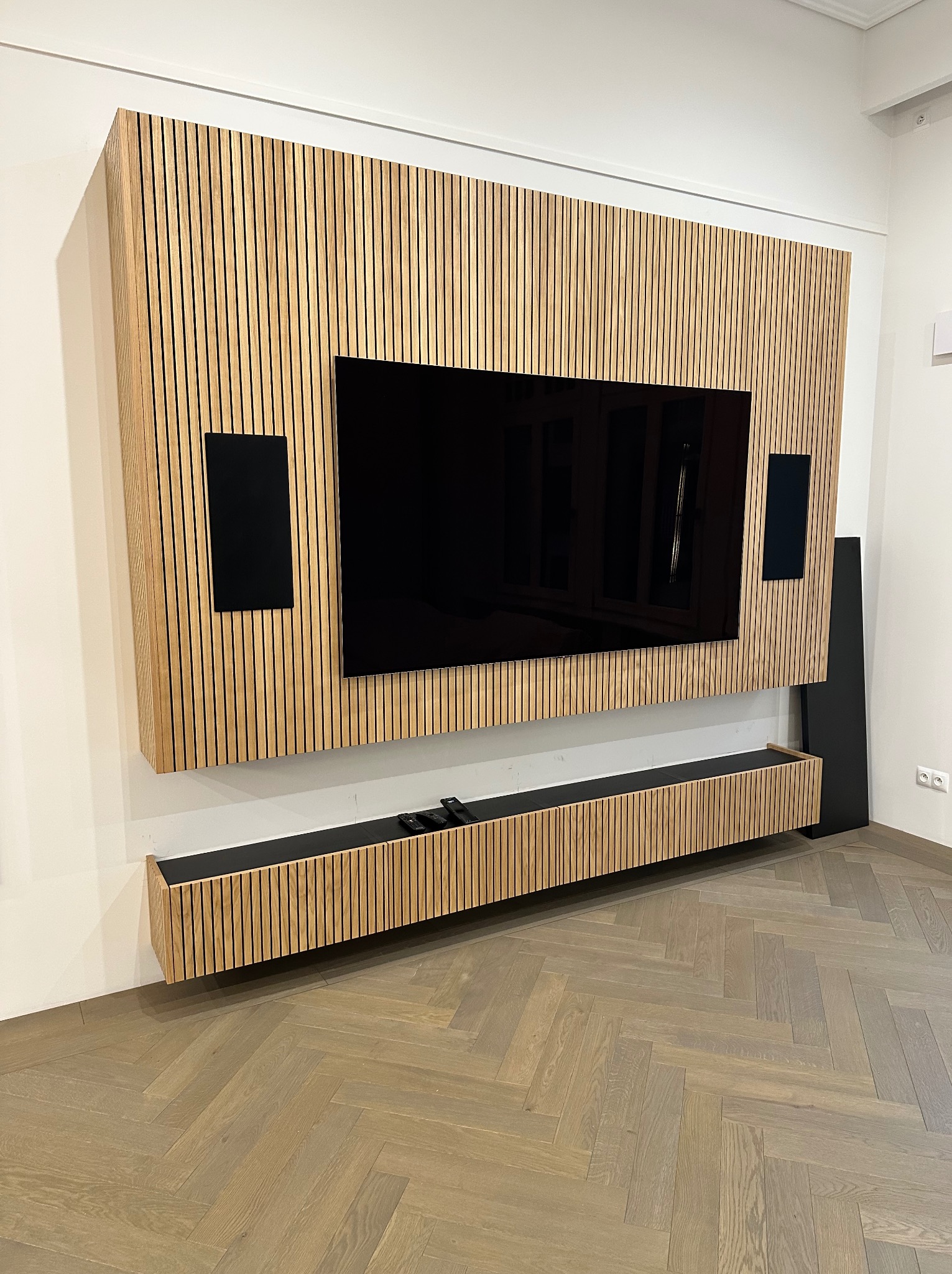 Drewniana zabudowa ściany z telewizorem i głośnikami, z listew w kolorze jasnego drewna, z szafką RTV na dole, parkiet ułożony w jodełkę.