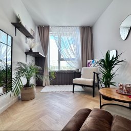 VibeHome Aleksandra Syktus - Jasne, przestronne wnętrze pokoju dziennego z parkietem w jodełkę, dużym oknem z zasłonami, lustrem w czarnej ramie, roślinami doniczkowymi i minimalistycznym umeblowaniem.