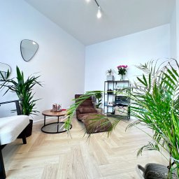 VibeHome Aleksandra Syktus - Jasne, minimalistyczne wnętrze z parkietem w jodełkę, dwoma asymetrycznymi lustrami na ścianie, brązowym leżakiem i roślinami doniczkowymi.
