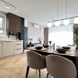 VibeHome Aleksandra Syktus - Nowoczesna kuchnia z jadalnią: beżowe szafki kuchenne, drewniany parkiet w jodełkę, stół z nakryciem, designerska lampa wisząca i dekoracje roślinne.