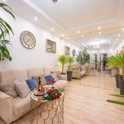 VibeHome Aleksandra Syktus - Jasny salon z beżową sofą, złotym stolikiem kawowym z dekoracją świąteczną i licznymi roślinami doniczkowymi, odbity w lustrzanej ścianie.