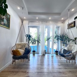 VibeHome Aleksandra Syktus - Jasne wnętrze z dwoma wiszącymi fotelami typu kokon, dekorowane poduszkami i futrzanymi narzutami, z parkietem i roślinami doniczkowymi, widok na taras przez przeszklone drzwi balkonowe.