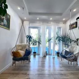 Jasne wnętrze z dwoma wiszącymi fotelami typu kokon, dekorowane poduszkami i futrzanymi narzutami, z parkietem i roślinami doniczkowymi, widok na taras przez przeszklone drzwi balkonowe.