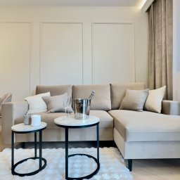 VibeHome Aleksandra Syktus - Beżowy narożnik z poduszkami, stoliki kawowe z szampanem, kwiaty i roślina doniczkowa w jasnym salonie z panelami ściennymi i drewnianą podłogą.