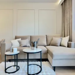 Beżowy narożnik z poduszkami, stoliki kawowe z szampanem, kwiaty i roślina doniczkowa w jasnym salonie z panelami ściennymi i drewnianą podłogą.