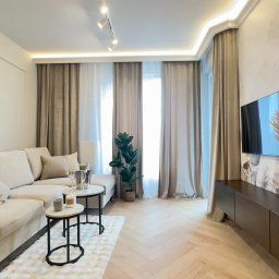 VibeHome Aleksandra Syktus - Jasne, przestronne wnętrze salonu z beżową sofą, stolikami kawowymi i telewizorem na ścianie z motywem roślinnym. Parkiet w jodełkę, dekoracyjne zasłony i subtelne oświetlenie.