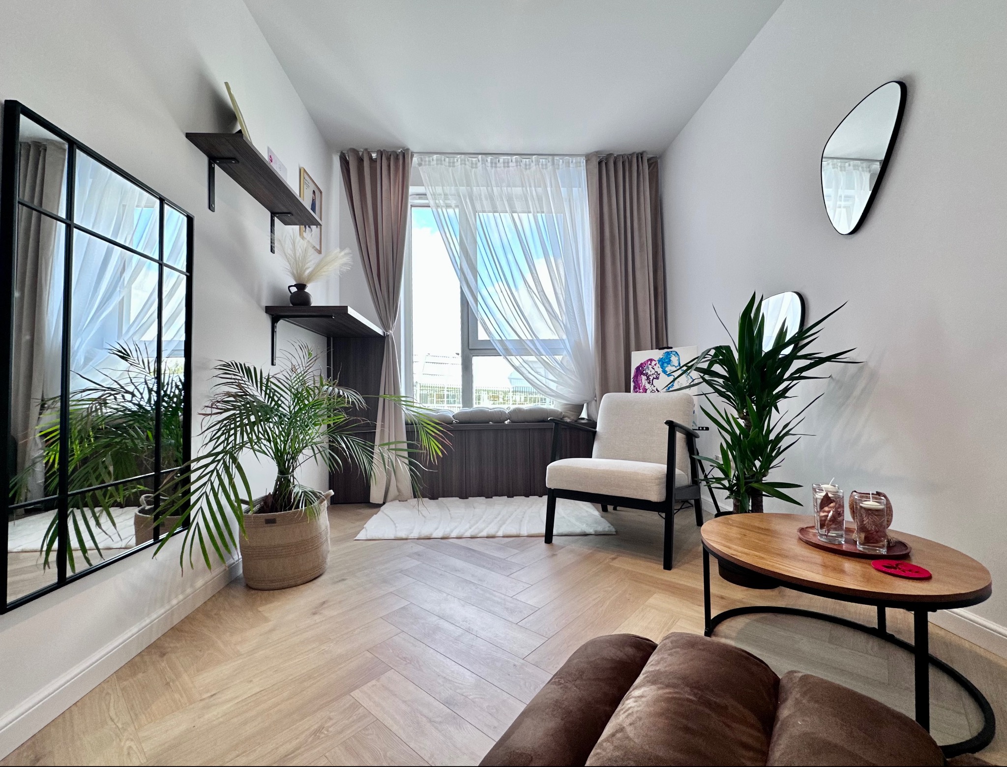 Jasne, przestronne wnętrze pokoju dziennego z parkietem w jodełkę, dużym oknem z zasłonami, lustrem w czarnej ramie, roślinami doniczkowymi i minimalistycznym umeblowaniem.