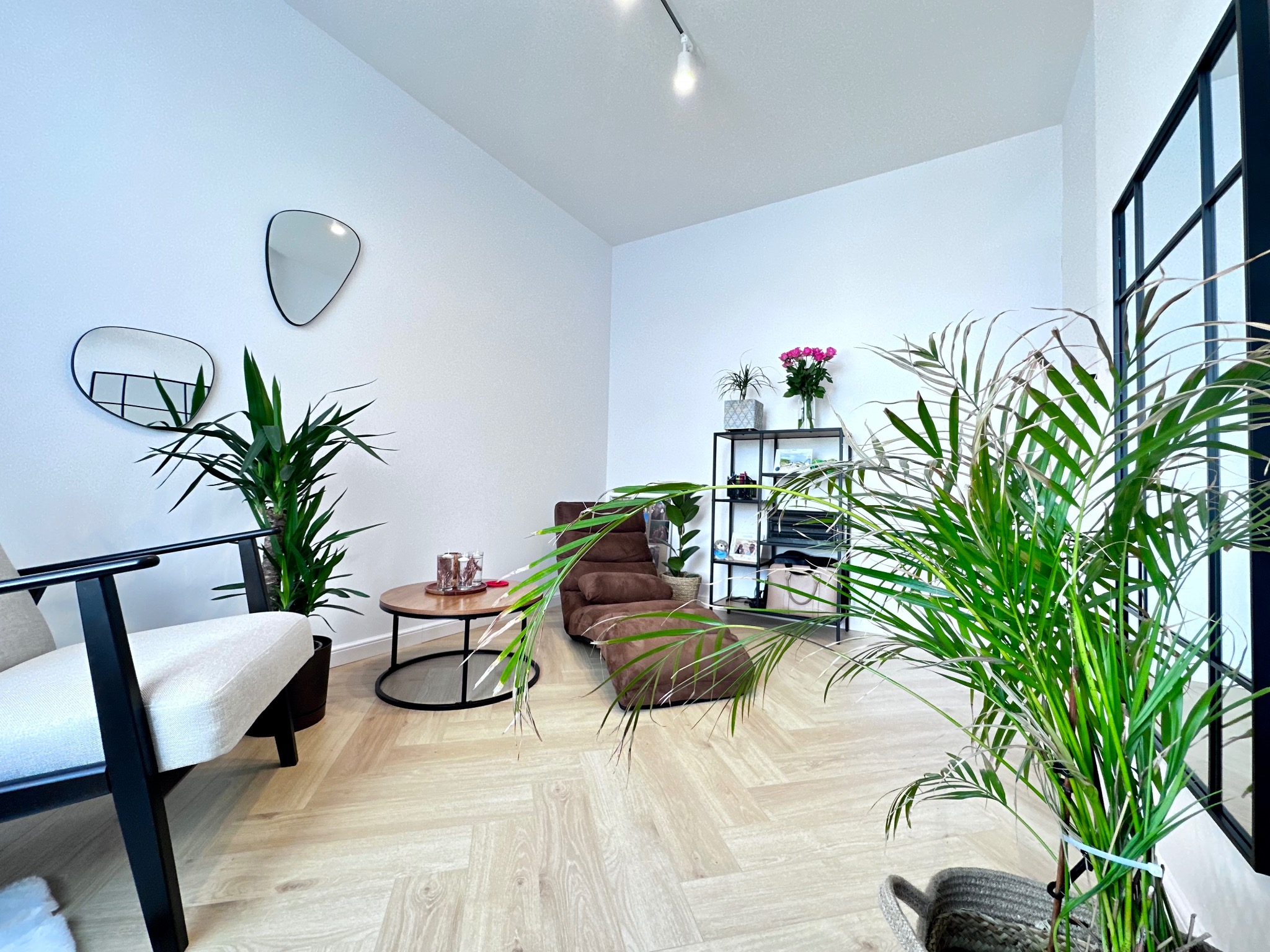 Jasne, minimalistyczne wnętrze z parkietem w jodełkę, dwoma asymetrycznymi lustrami na ścianie, brązowym leżakiem i roślinami doniczkowymi.