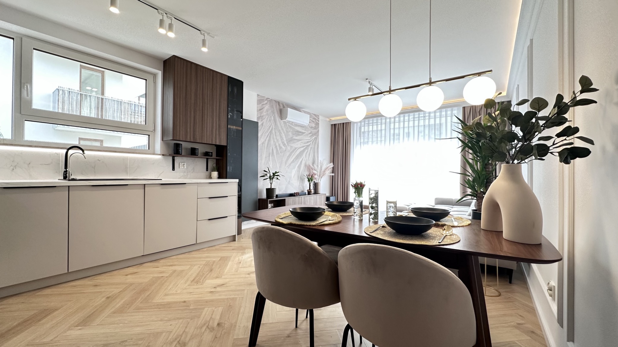 Nowoczesna kuchnia z jadalnią: beżowe szafki kuchenne, drewniany parkiet w jodełkę, stół z nakryciem, designerska lampa wisząca i dekoracje roślinne.