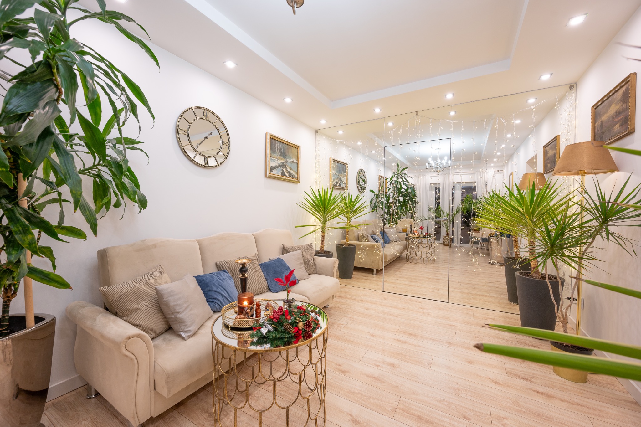 Jasny salon z beżową sofą, złotym stolikiem kawowym z dekoracją świąteczną i licznymi roślinami doniczkowymi, odbity w lustrzanej ścianie.