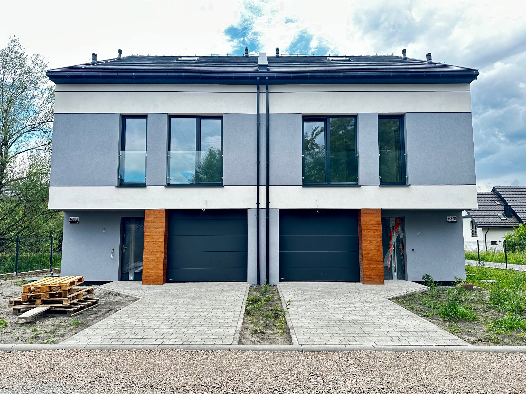 Dwukondygnacyjny, bliźniaczy dom w nowoczesnym stylu z płaskim dachem, szarą elewacją i drewnianymi akcentami, z czarnymi bramami garażowymi i paletami przed wejściem do jednego z segmentów.